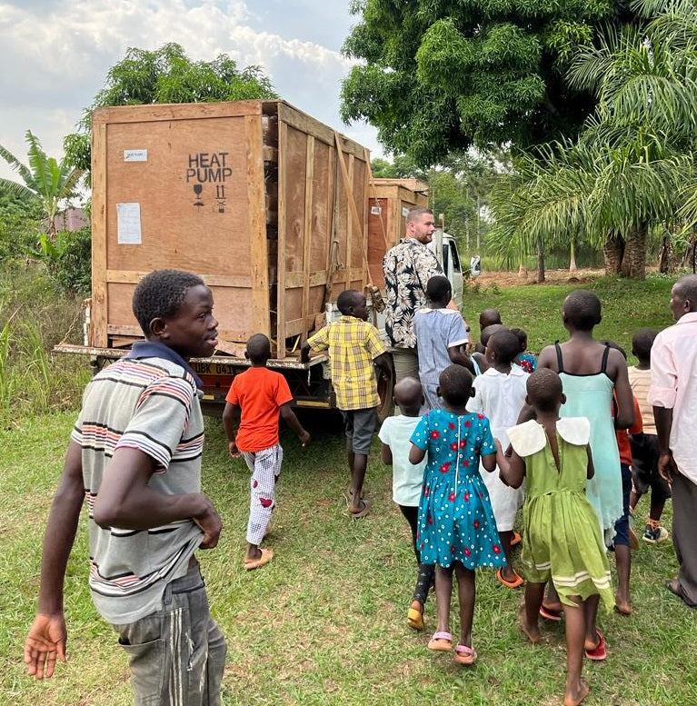 Ugandan village children follow a truck with big wooden boxes on through a lush green environment with trees.